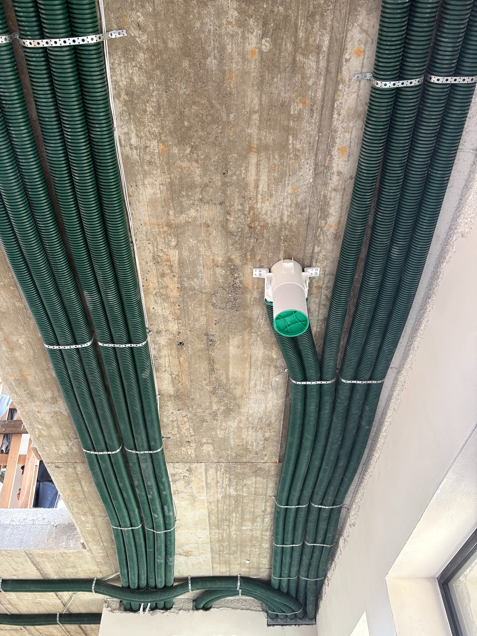 Zielone, karbowane rury wentylacyjne zamocowane do surowego betonowego stropu za pomocą metalowych obejm. Widoczny biały rozdzielacz z zielonym elementem.