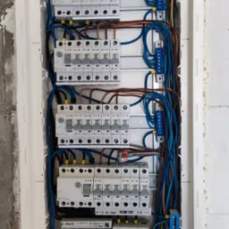 Rozdzielnia elektryczna z zamontowanymi bezpiecznikami EATON, kable w listwie, widok z bliska na instalację w białej obudowie na tle szarej ściany.