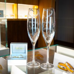AMORO SPÓŁKA Z OGRANICZONĄ ODPOWIEDZIALNOŚCIĄ - Dwa spersonalizowane kieliszki do szampana z imionami i datą, obok pudełko z obrączkami, na szklanej powierzchni ekspozycyjnej w sklepie jubilerskim.