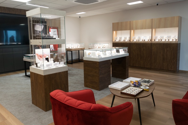 Wnętrze salonu jubilerskiego z ekspozytorami biżuterii, stolikiem z próbkami i czerwonymi fotelami, tworzące elegancką przestrzeń handlową.