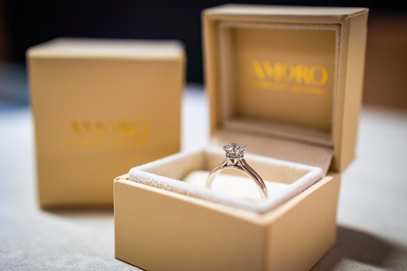 Elegancki pierścionek zaręczynowy z brylantem w otwartym, kremowym pudełku marki AMORO. W tle drugie, zamknięte pudełko. Subtelne oświetlenie podkreśla blask kamienia.