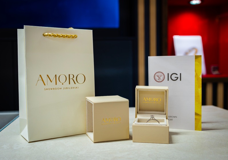 Elegancki zestaw prezentowy z pierścionkiem od jubilera Amoro, obejmujący pudełko, torbę i certyfikat IGI, na neutralnym tle.