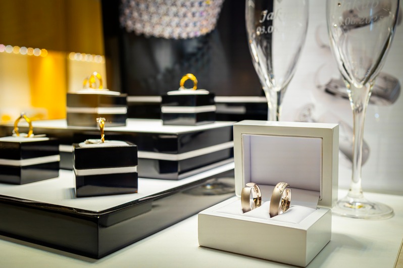 Zestaw złotych obrączek w białym pudełku na tle ekspozycji z pierścionkami i kieliszkami z grawerem. Elegancki upominek ślubny, idealny na rocznicę.