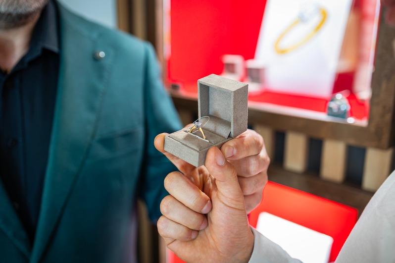 Mężczyzna prezentuje pierścionek zaręczynowy w szarym pudełku na tle ekspozycji biżuterii w sklepie. Elegancki garnitur i dbałość o detale podkreślają wyjątkowość momentu.