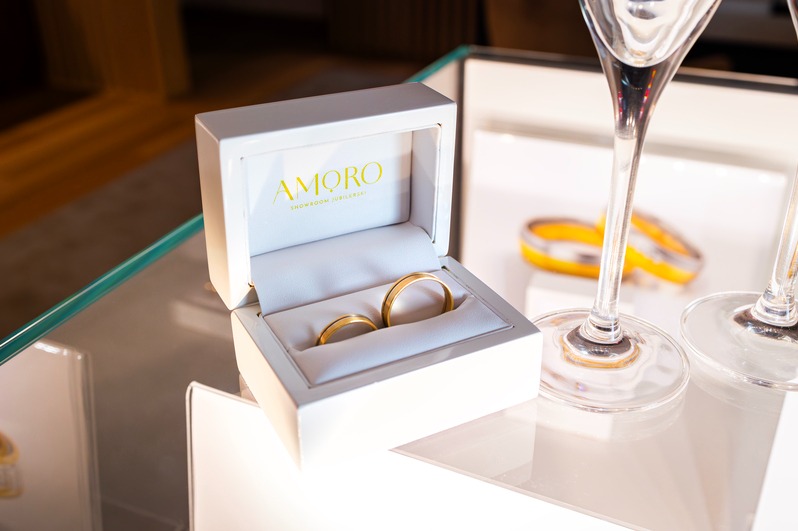 Złote obrączki w białym pudełku marki AMORO, ustawione na szklanym blacie obok kieliszków. Elegancki detal jubilerski na ślub, z bliska.