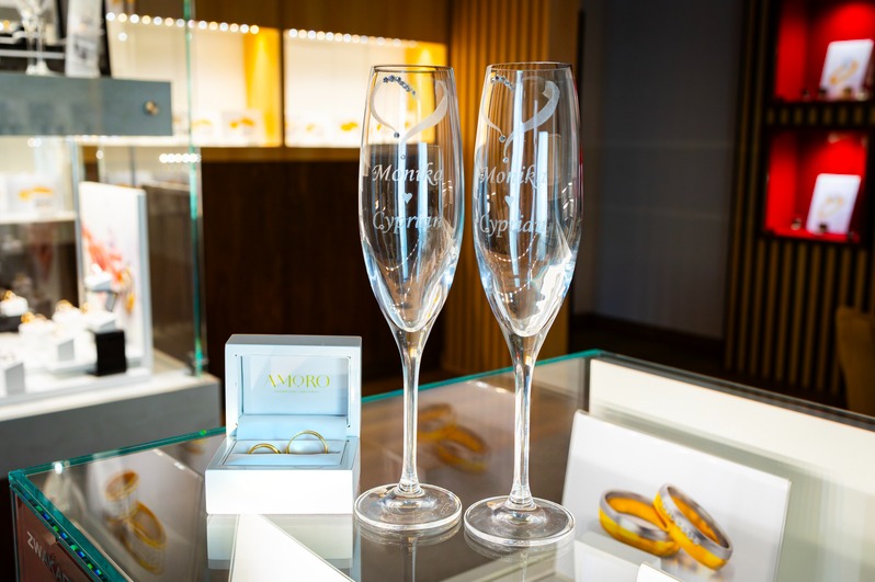 Dwa spersonalizowane kieliszki do szampana z imionami i datą, obok pudełko z obrączkami, na szklanej powierzchni ekspozycyjnej w sklepie jubilerskim.