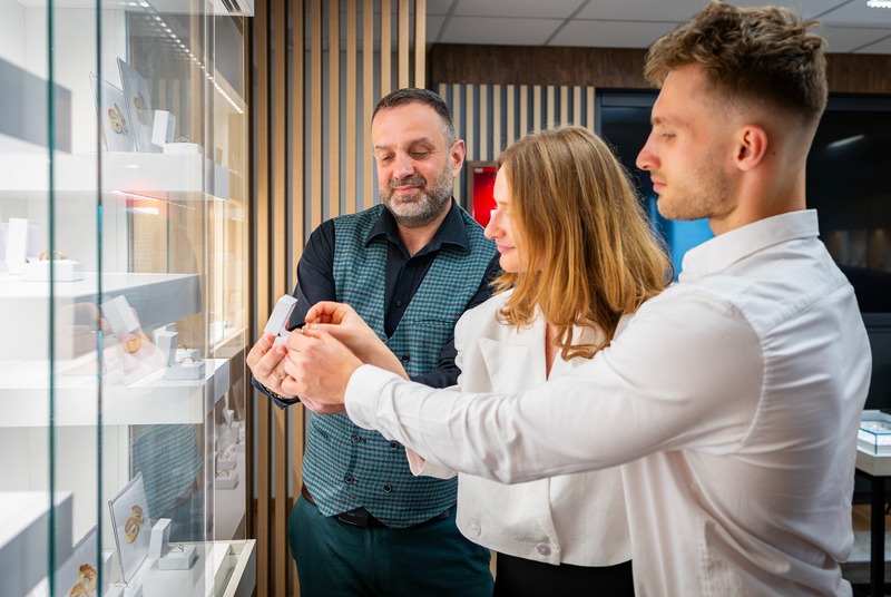 Kobieta i mężczyzna oglądają pierścionek zaręczynowy w salonie jubilerskim, doradca prezentuje detal. Szklana witryna z biżuterią w tle.