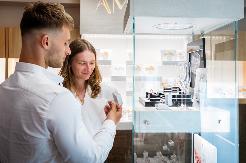 Para ogląda pierścionek zaręczynowy w eleganckim salonie jubilerskim z oświetloną witryną pełną biżuterii. Skupienie na detalu i wyborze idealnego prezentu.