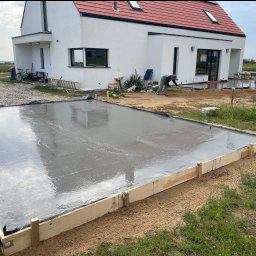 FUNDKOP SP Z O.O. - Wylany fundament pod budowę domu jednorodzinnego. Widoczny szalunek z desek i gładka powierzchnia świeżego betonu. W tle dom w stylu nowoczesnej stodoły i koparka.
