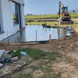 FUNDKOP SP Z O.O. - Wylany beton pod taras przy domu jednorodzinnym. Żółta koparka Volvo w tle. Widoczne drewniane szalunki i folia. Butelka wody na ziemi. Częściowo pochmurne niebo.