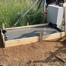 FUNDKOP SP Z O.O. - Wylany beton w szalunku z desek, zabezpieczony folią. Otoczenie to ziemia i trawa. W tle widoczny sprzęt budowlany i ogrodzenie z siatki.