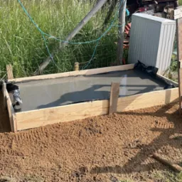 Wylany beton w szalunku z desek, zabezpieczony folią. Otoczenie to ziemia i trawa. W tle widoczny sprzęt budowlany i ogrodzenie z siatki.