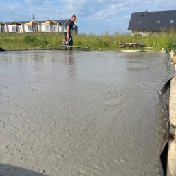 FUNDKOP SP Z O.O. - Wylewanie fundamentów: pracownik wyrównuje świeży beton na przygotowanym terenie budowy, w tle domy jednorodzinne i błękitne niebo.