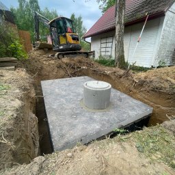FUNDKOP SP Z O.O. - Montaż betonowego szamba w wykopie; widoczny żółty mini-koparka Volvo na skraju wykopu i betonowy zbiornik z włazem. W tle budynek z czerwoną dachówką.