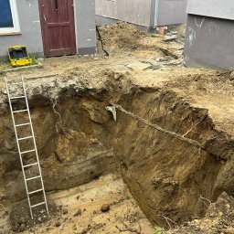 FUNDKOP SP Z O.O. - Głęboki wykop w ziemi, prawdopodobnie pod fundamenty, z widoczną drabiną i żółtą skrzynką narzędziową obok budynku. Warstwowa struktura ziemi w wykopie.