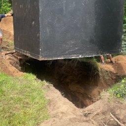 FUNDKOP SP Z O.O. - Czarny, betonowy szambonurek zawieszony nad wykopem w ziemi, wspierany przez osoby po bokach. Widoczne trawa i drzewa w tle. Etap instalacji szamba.