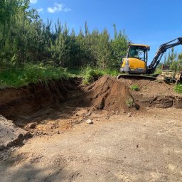 FUNDKOP SP Z O.O. - Żółta koparka Volvo na placu budowy, załadowuje ziemię do pomarańczowej ciężarówki. Wykop w ziemi, zielone drzewa w tle, błękitne niebo.