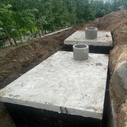 FUNDKOP SP Z O.O. - Montaż dwóch betonowych szamb z kominkami w wykopie, widok z góry. Prace ziemne w Kartuzach. Szamba z betonu, grunt, zieleń w tle.