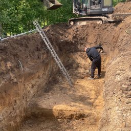 FUNDKOP SP Z O.O. - Głęboki wykop w ziemi z drabiną, pracownikiem z łopatą i koparką ECR50D na górze. Widok na prace ziemne w słoneczny dzień.