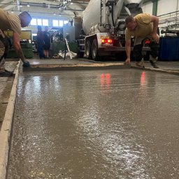 FUNDKOP SP Z O.O. - Wylewka betonowa wyrównywana przez dwóch robotników. W tle betoniarka i obserwujący. Widoczne światła stop betoniarki, szalunek i mokra powierzchnia betonu.