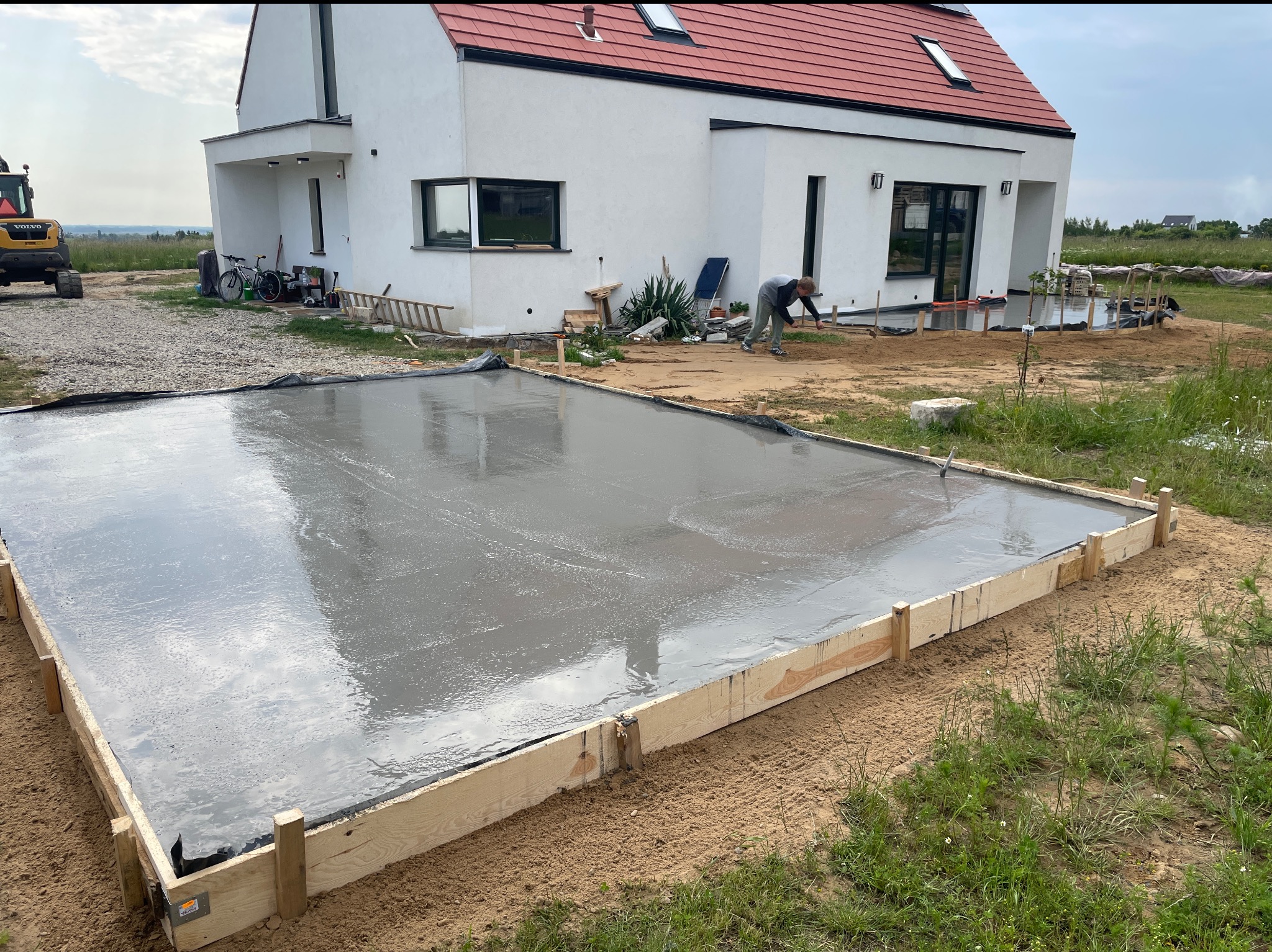 Wylany fundament pod budowę domu jednorodzinnego. Widoczny szalunek z desek i gładka powierzchnia świeżego betonu. W tle dom w stylu nowoczesnej stodoły i koparka.