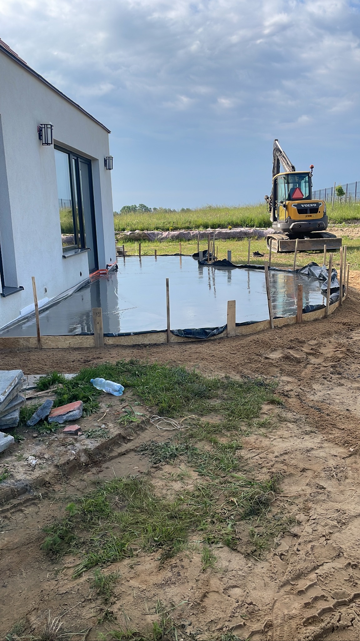Wylany beton pod taras przy domu jednorodzinnym. Żółta koparka Volvo w tle. Widoczne drewniane szalunki i folia. Butelka wody na ziemi. Częściowo pochmurne niebo.