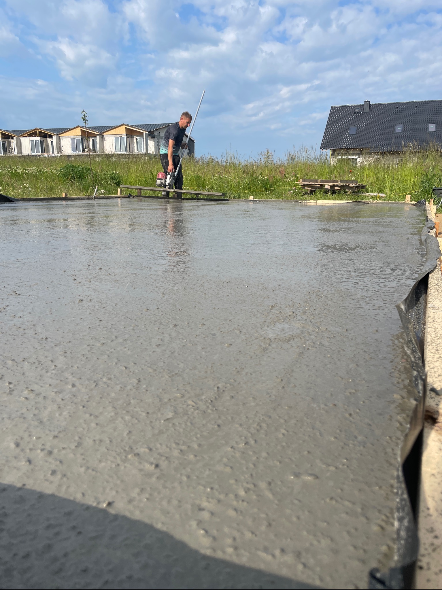 Wylewanie fundamentów: pracownik wyrównuje świeży beton na przygotowanym terenie budowy, w tle domy jednorodzinne i błękitne niebo.