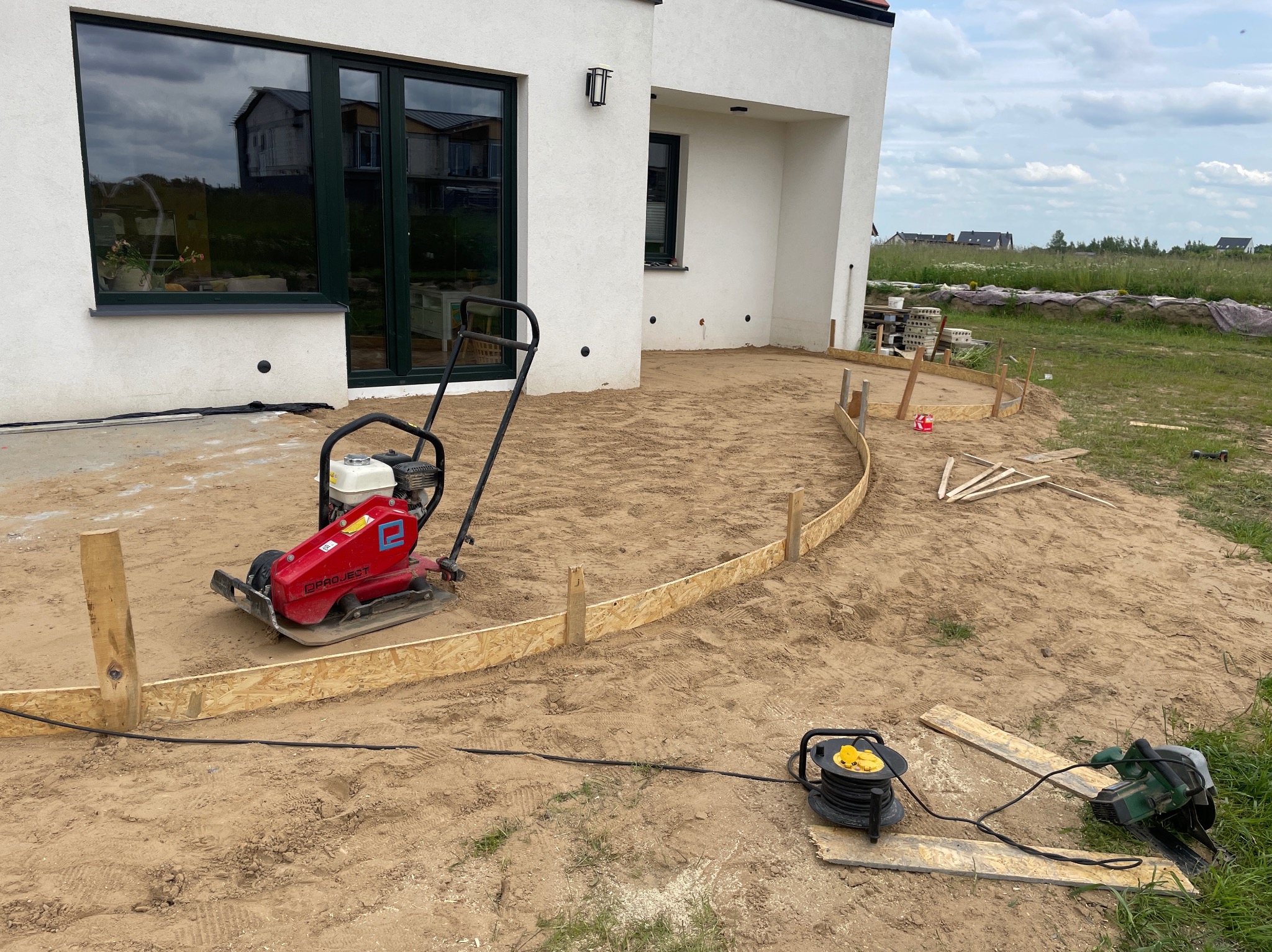 Przygotowanie podłoża pod taras: ubijarka spalinowa, obrzeża z desek, piasek. Widoczny dom z dużymi oknami i trawa. Prace ziemne w toku.