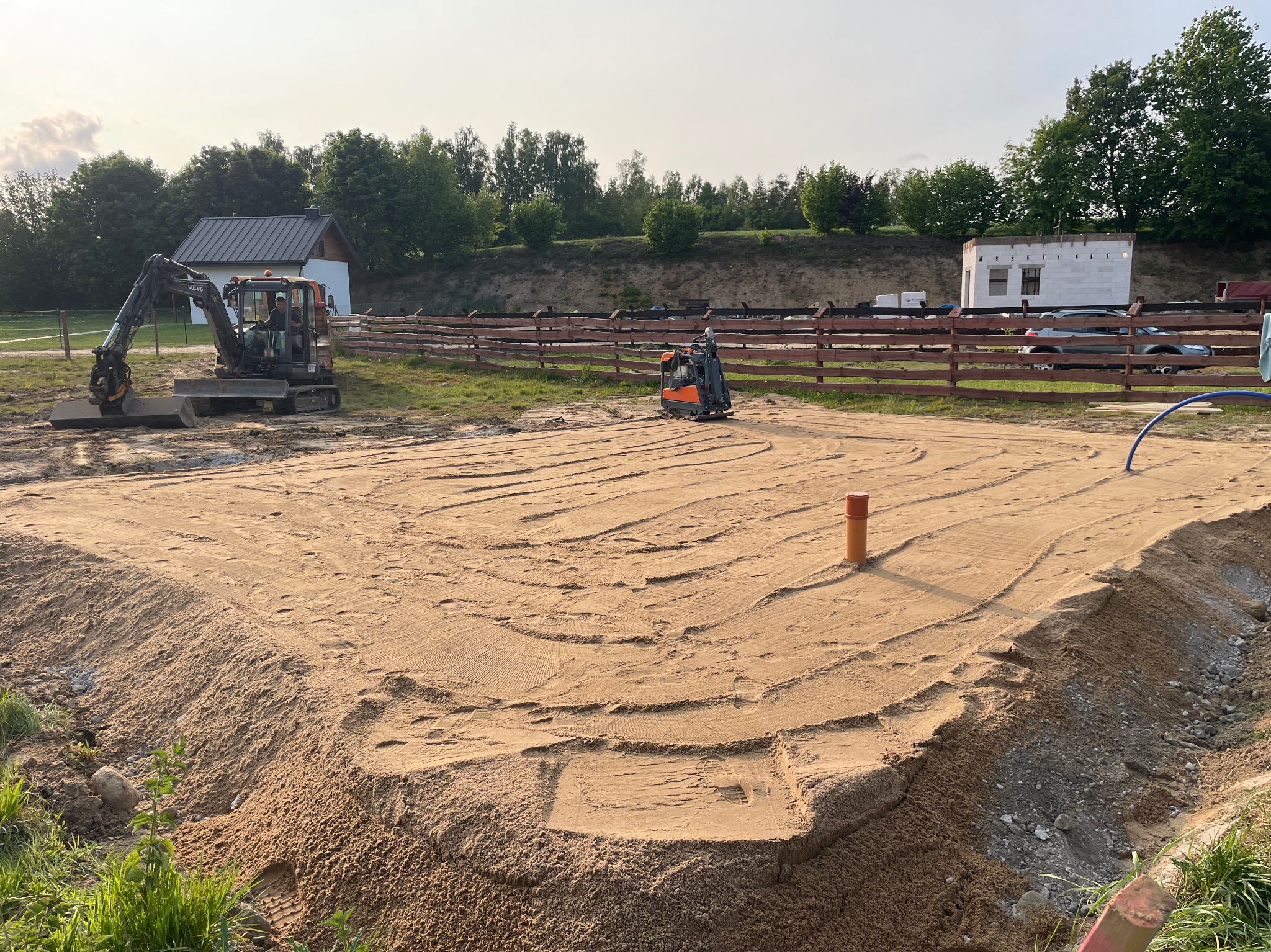 Niwelacja terenu pod budowę z użyciem minikoparki i ubijarki. Żółty piasek, wyrównany teren, w tle budynek w trakcie budowy i drzewa.