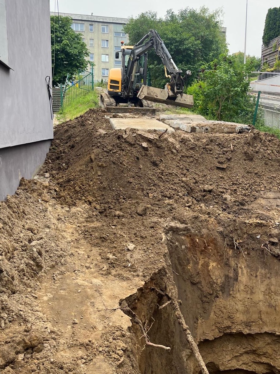 Żółta koparka Volvo usuwa betonowe płyty obok wykopu ziemnego. W tle budynek mieszkalny i zieleń. Prace ziemne w toku.