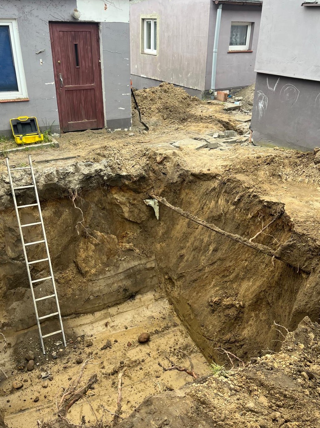 Głęboki wykop w ziemi, prawdopodobnie pod fundamenty, z widoczną drabiną i żółtą skrzynką narzędziową obok budynku. Warstwowa struktura ziemi w wykopie.