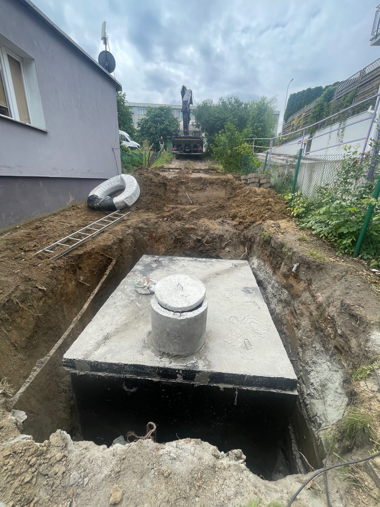 Betonowy zbiornik w wykopie, widoczna drabina i zwój rur. W tle koparka i budynek. Prace ziemne w toku, instalacja zbiornika.