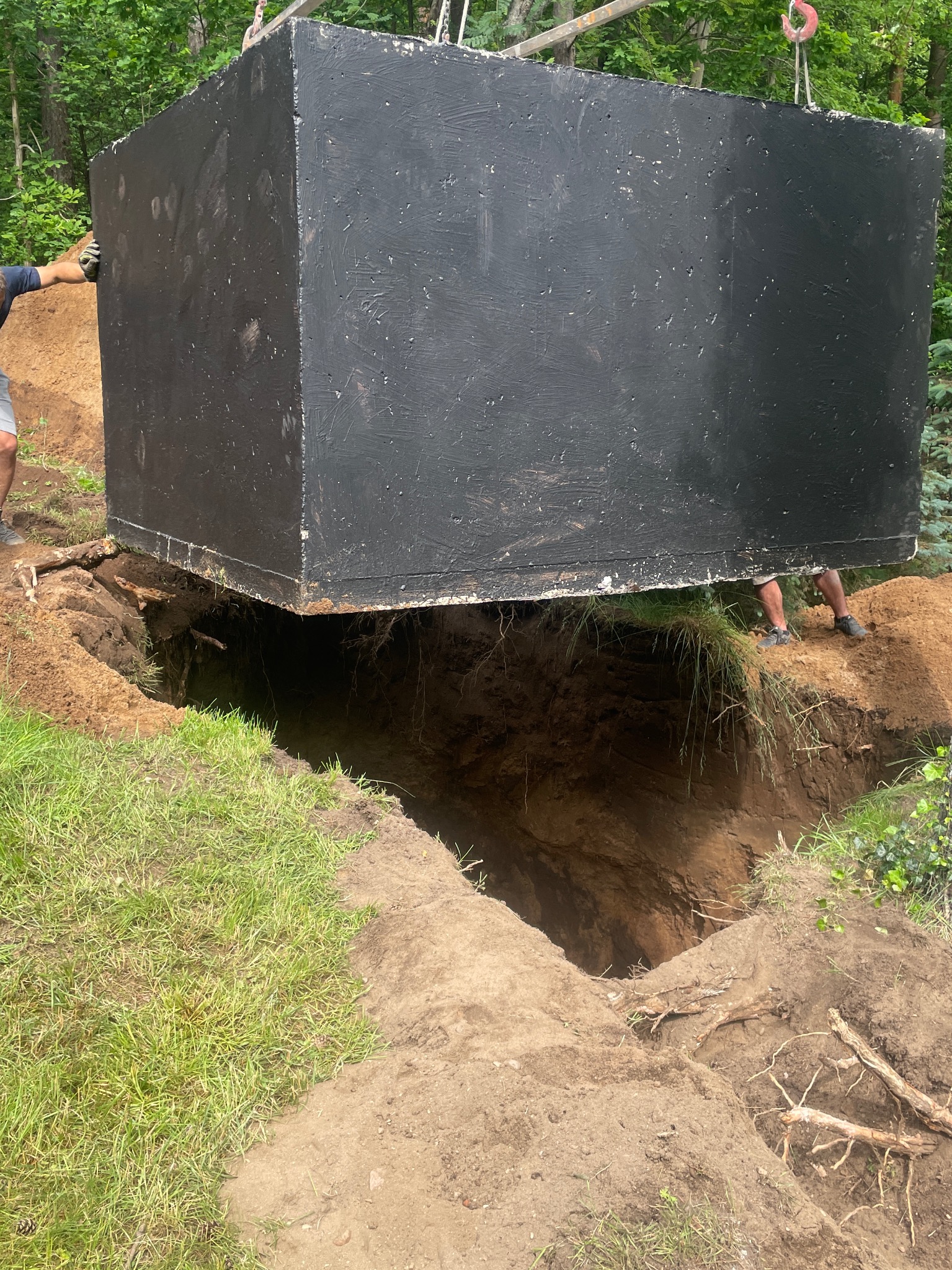 Czarny, betonowy szambonurek zawieszony nad wykopem w ziemi, wspierany przez osoby po bokach. Widoczne trawa i drzewa w tle. Etap instalacji szamba.