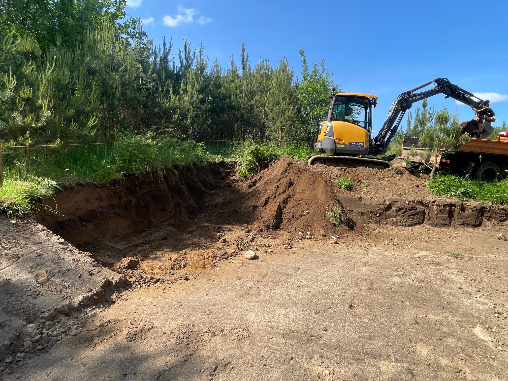 Żółta koparka Volvo na placu budowy, załadowuje ziemię do pomarańczowej ciężarówki. Wykop w ziemi, zielone drzewa w tle, błękitne niebo.
