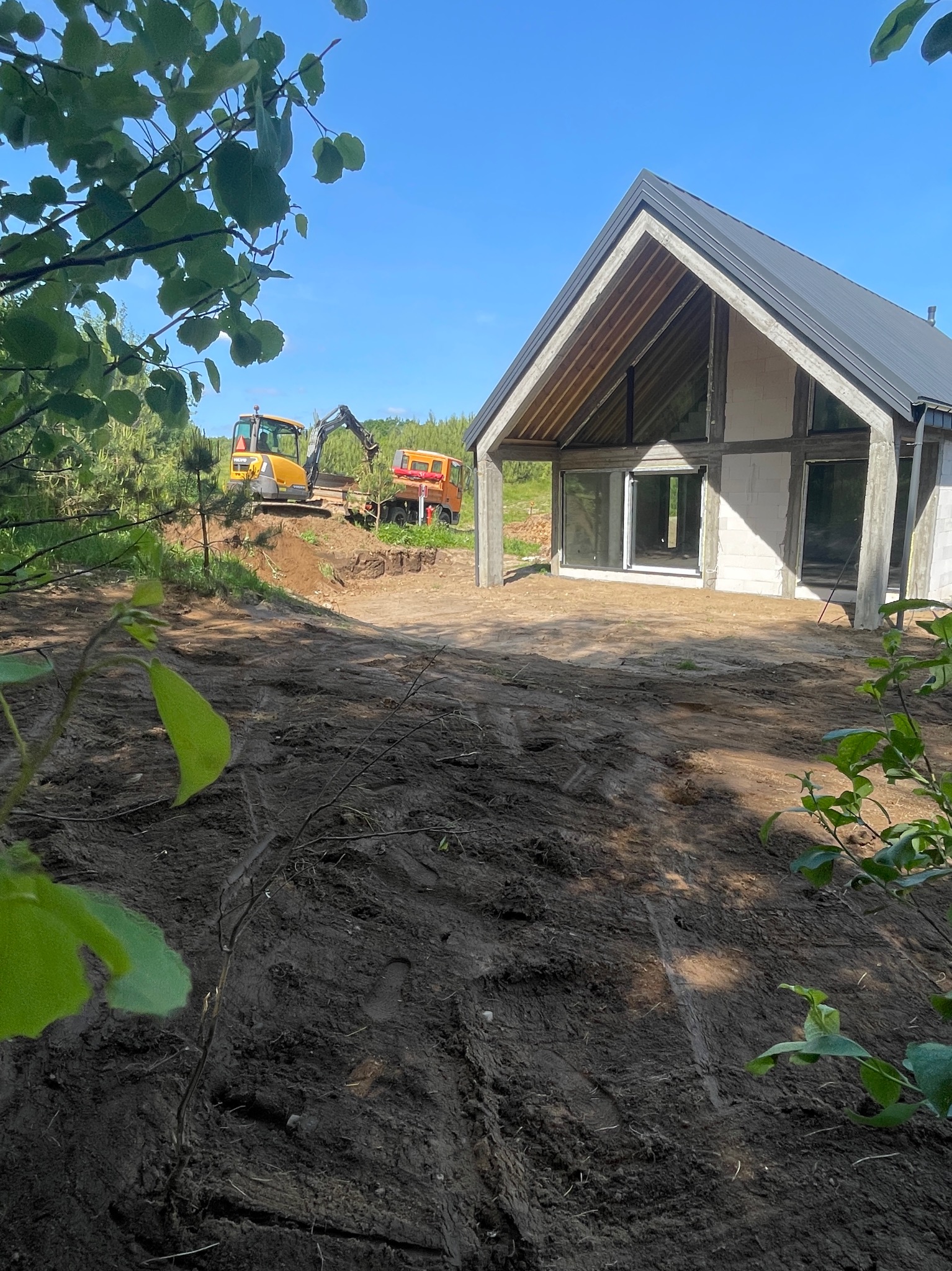 Budowa domu w trakcie: surowy budynek z oknami, w tle koparka i wywrotka na nierównym terenie. Ujęcie z dołu, widoczne liście drzew.