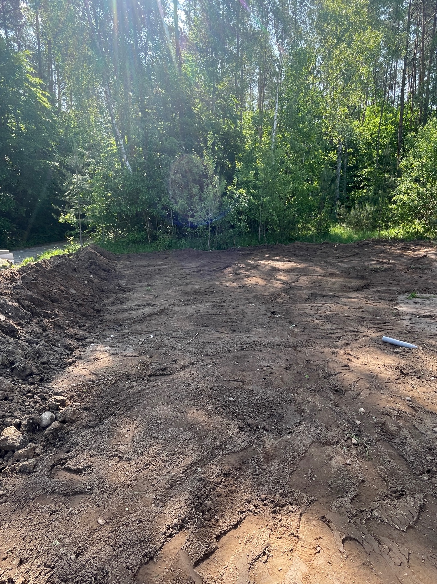 Przygotowany teren pod budowę, wyrównana ziemia z widocznymi śladami opon, otoczona zielenią drzew. W tle las, jasne światło słoneczne.