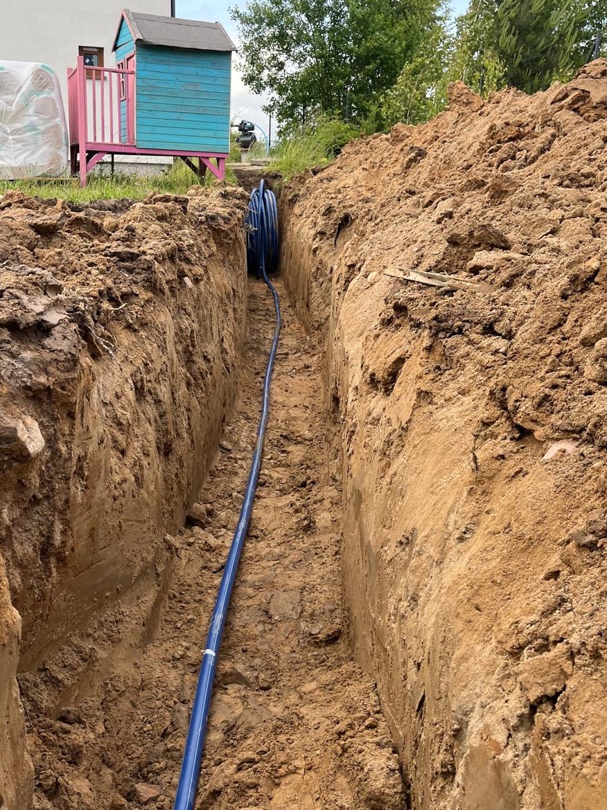 Wąski wykop w ziemi z niebieską rurą wewnątrz, prowadzącą w głąb. Widoczny fragment domku dziecięcego i roślinność w tle. Ujęcie z góry, podkreślające głębokość wykopu.