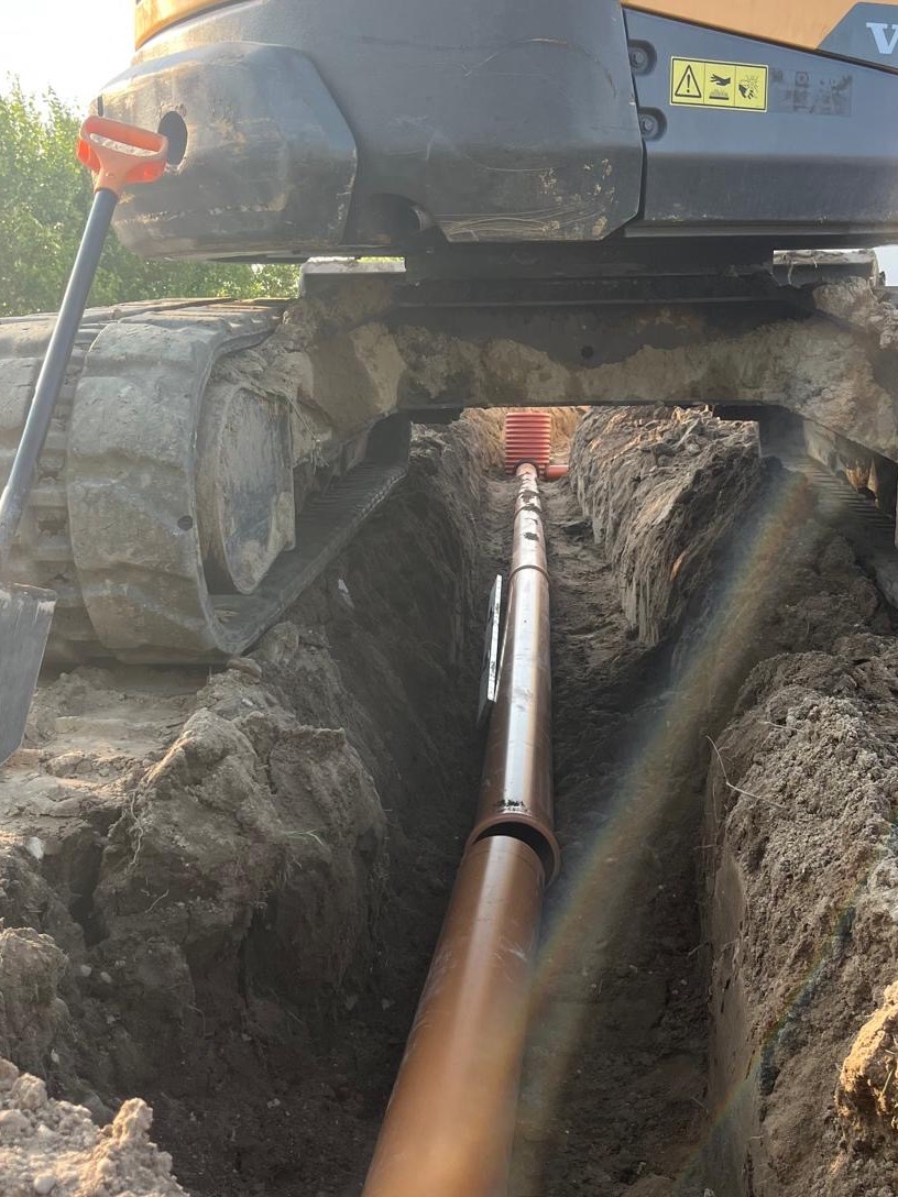 Brązowa rura ułożona w wykopie, widoczna spod spodu koparki. Pomarańczowa rura kanalizacyjna w tle. Praca ziemna w toku, częściowo oświetlona słońcem.