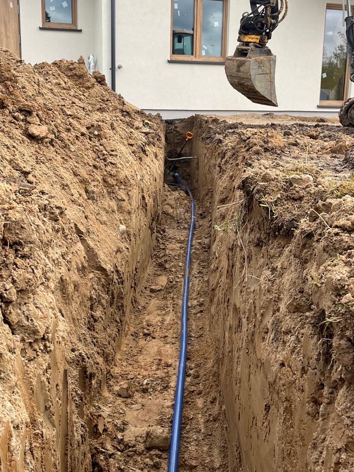 Wykopany rów z niebieską rurą w ziemi na tle jasnego budynku z oknami. Nad rowem widoczna łyżka koparki. Etap prac ziemnych.