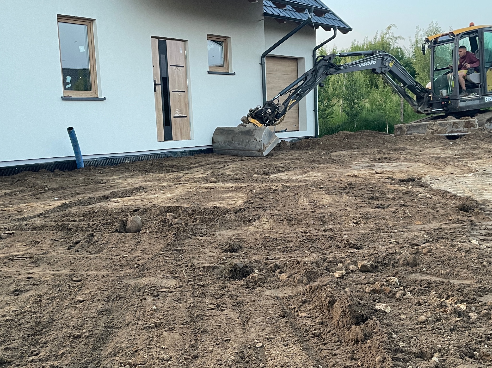 Żółta koparka Volvo wyrównuje teren wokół jasnego domu z brązowymi oknami i drzwiami. Widoczny fragment niebieskiej rury. Roboty ziemne w toku.
