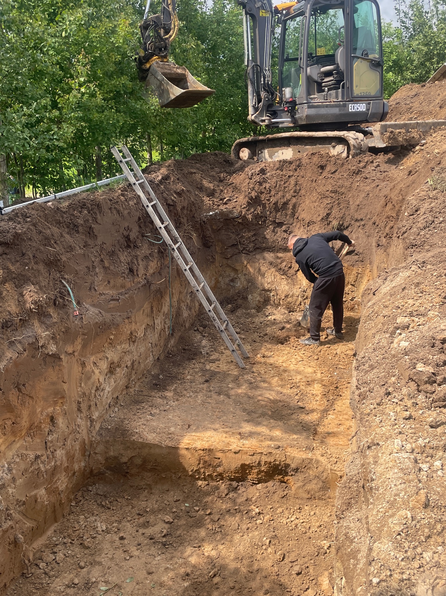Głęboki wykop w ziemi z drabiną, pracownikiem z łopatą i koparką ECR50D na górze. Widok na prace ziemne w słoneczny dzień.