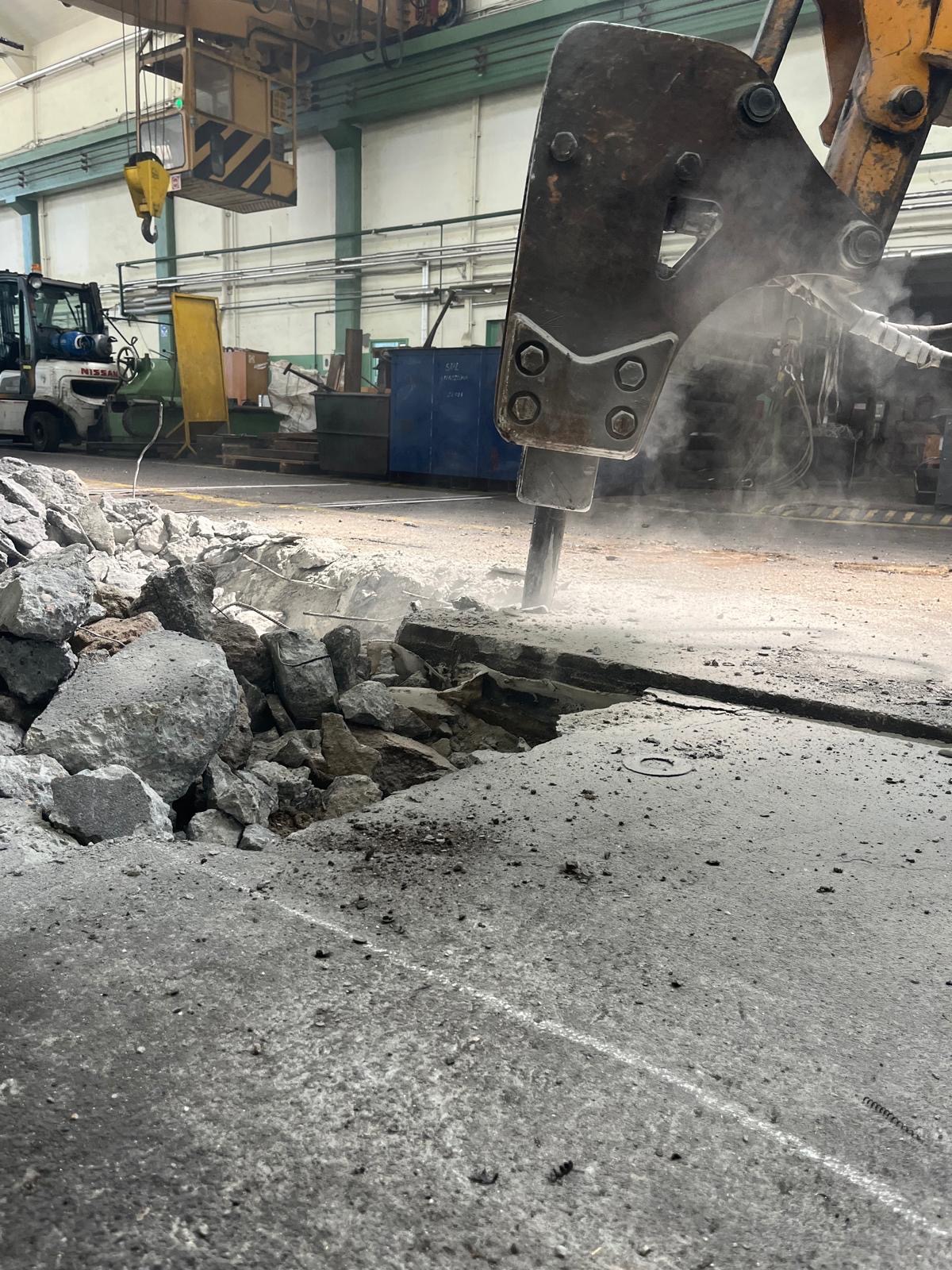 Młot pneumatyczny kruszy betonową posadzkę w hali przemysłowej. W tle widoczny wózek widłowy i elementy konstrukcji hali.