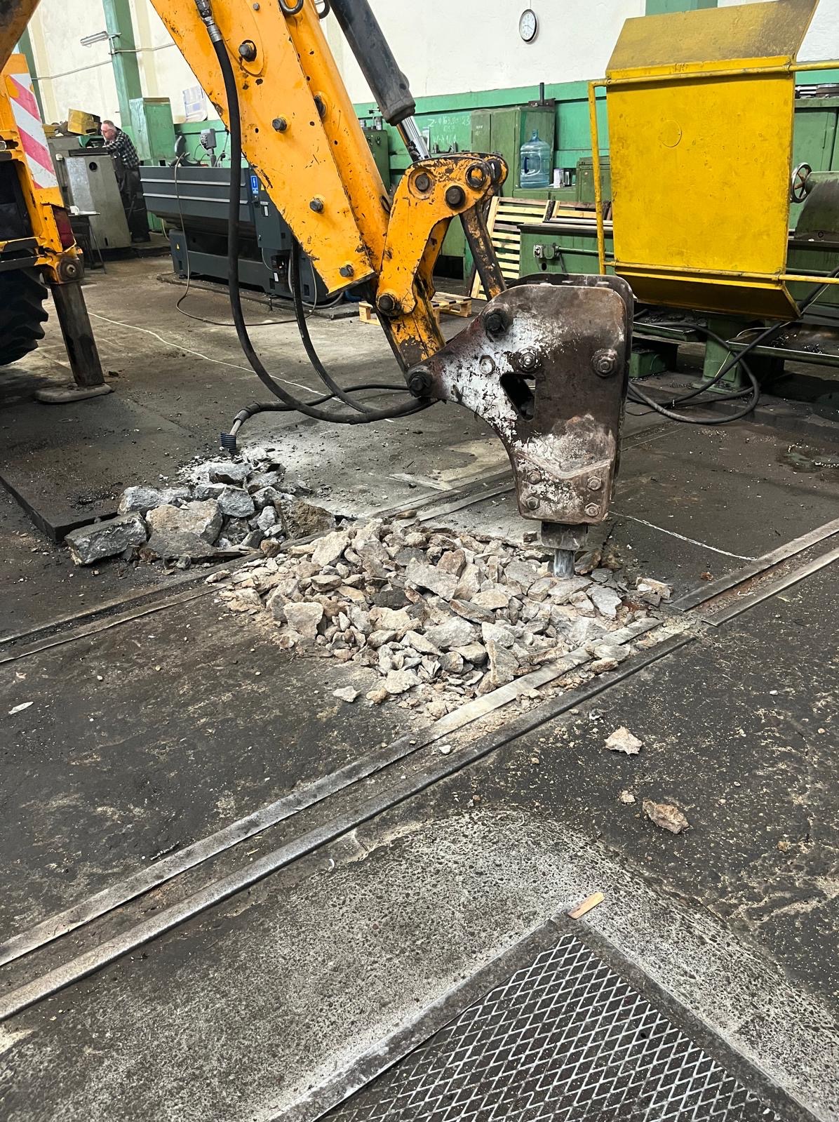 Żółty młot pneumatyczny kruszy beton na podłodze w hali. Widoczne metalowe szyny w posadzce. W tle maszyny i pracownik.
