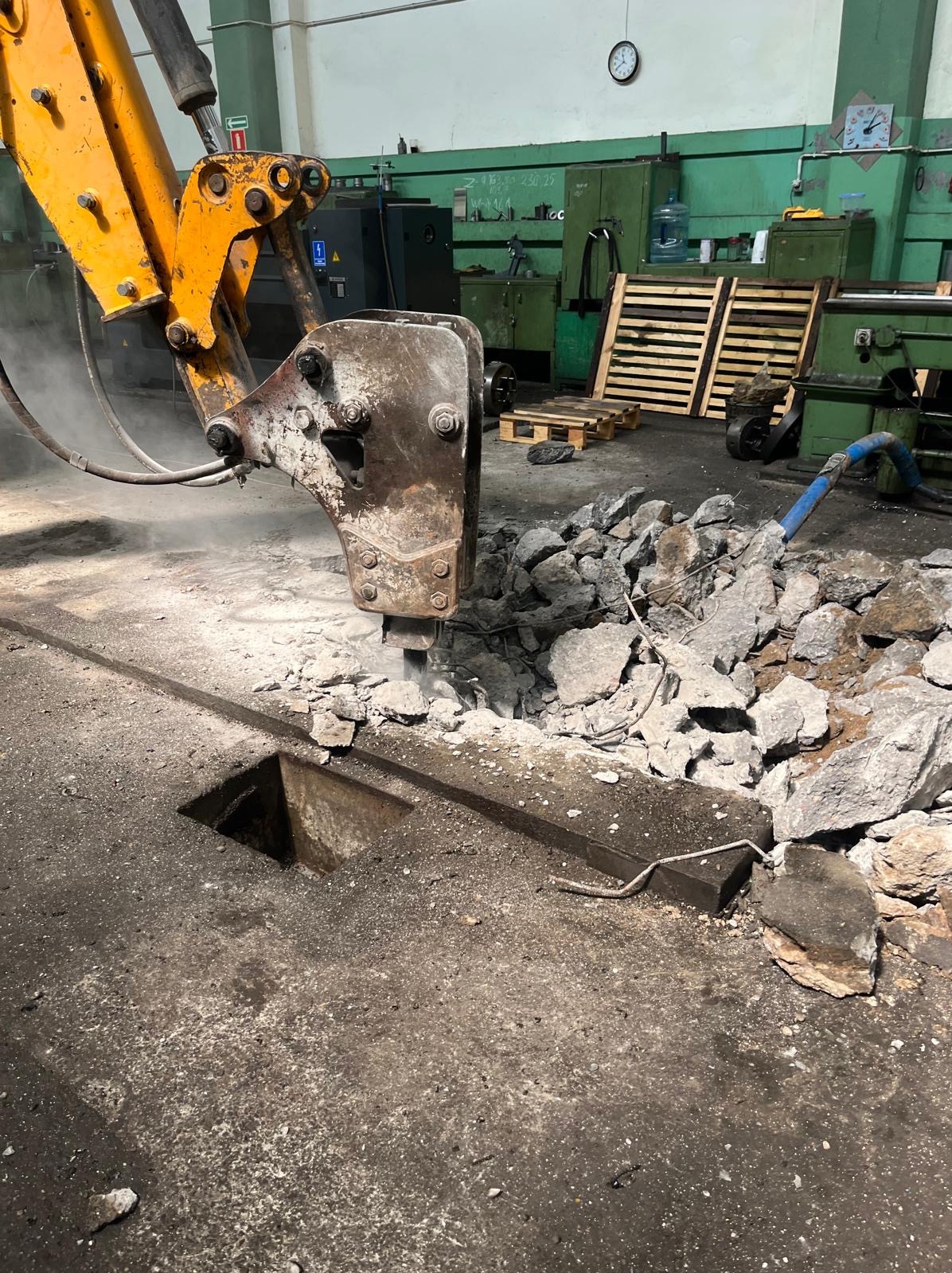 Hydrauliczny młot wyburzeniowy kruszy betonową posadzkę w hali. Widoczny wykop i gruz. Maszyna budowlana w trakcie pracy, w tle palety i maszyny.