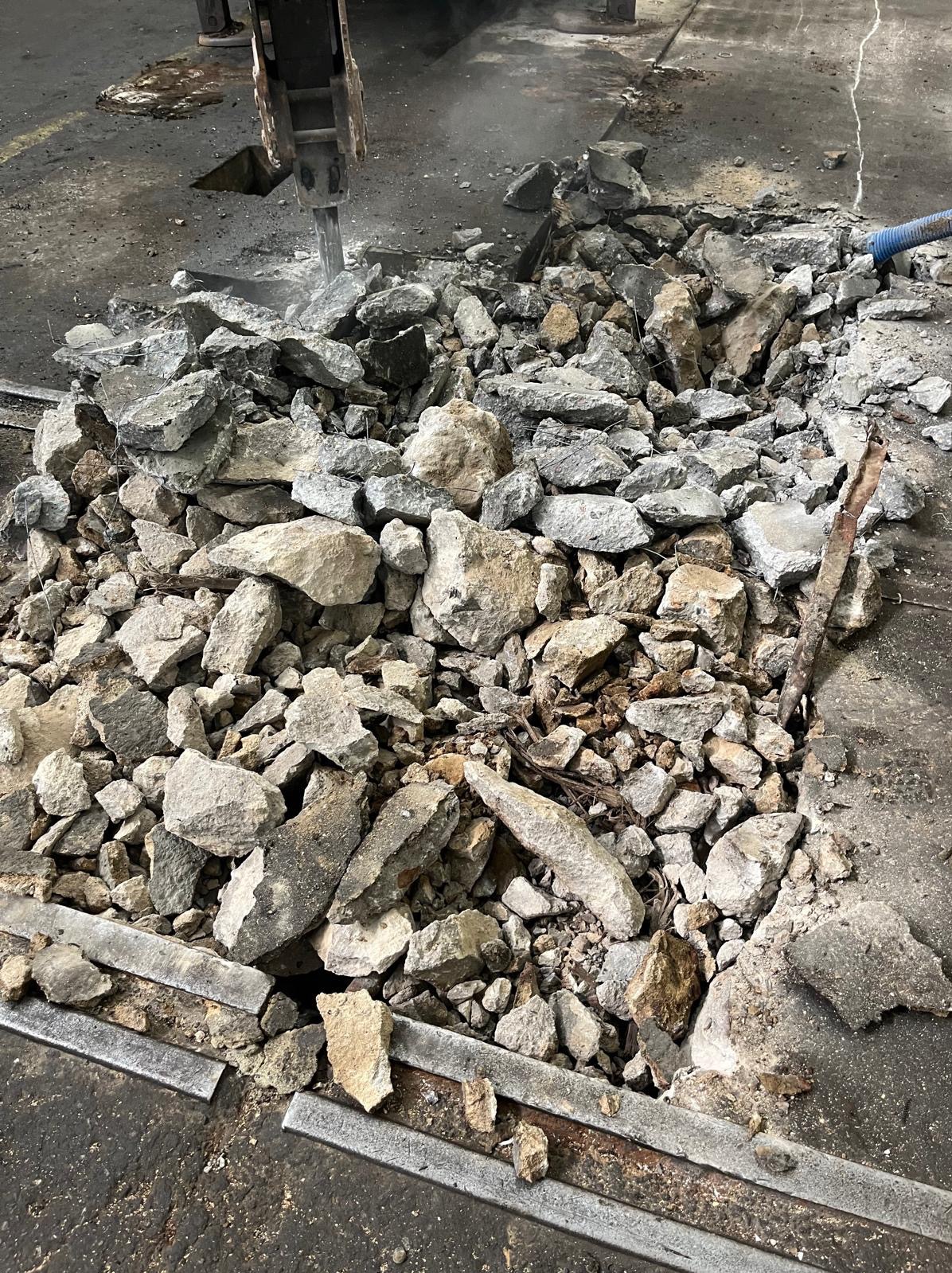 Rozbiórka posadzki betonowej młotem pneumatycznym. Fragment gruzu na podłożu z widocznymi szynami. Praca w toku, widoczne ślady pyłu.