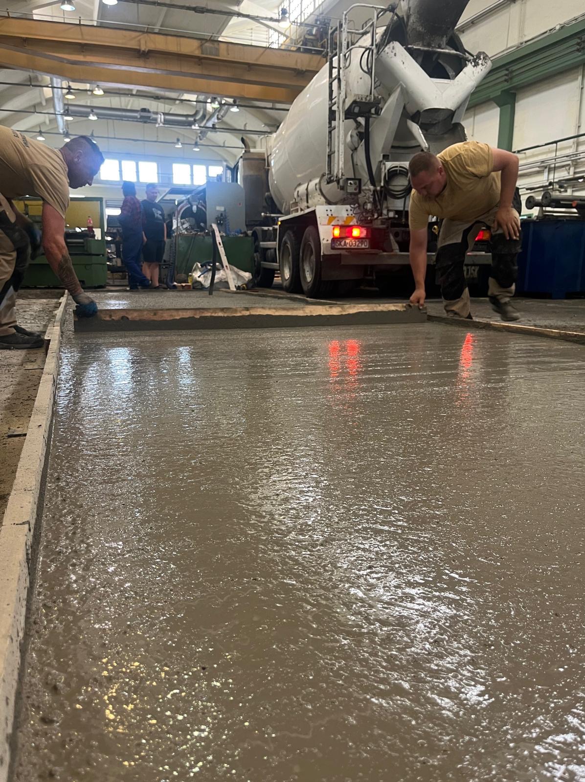 Wylewka betonowa wyrównywana przez dwóch robotników. W tle betoniarka i obserwujący. Widoczne światła stop betoniarki, szalunek i mokra powierzchnia betonu.