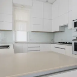 Biała, nowoczesna kuchnia z wyspą, jasnym blatem i sprzętem Bosch. Minimalistyczny design z przeszklonym panelem ściennym i subtelnymi zasłonami w oknie.