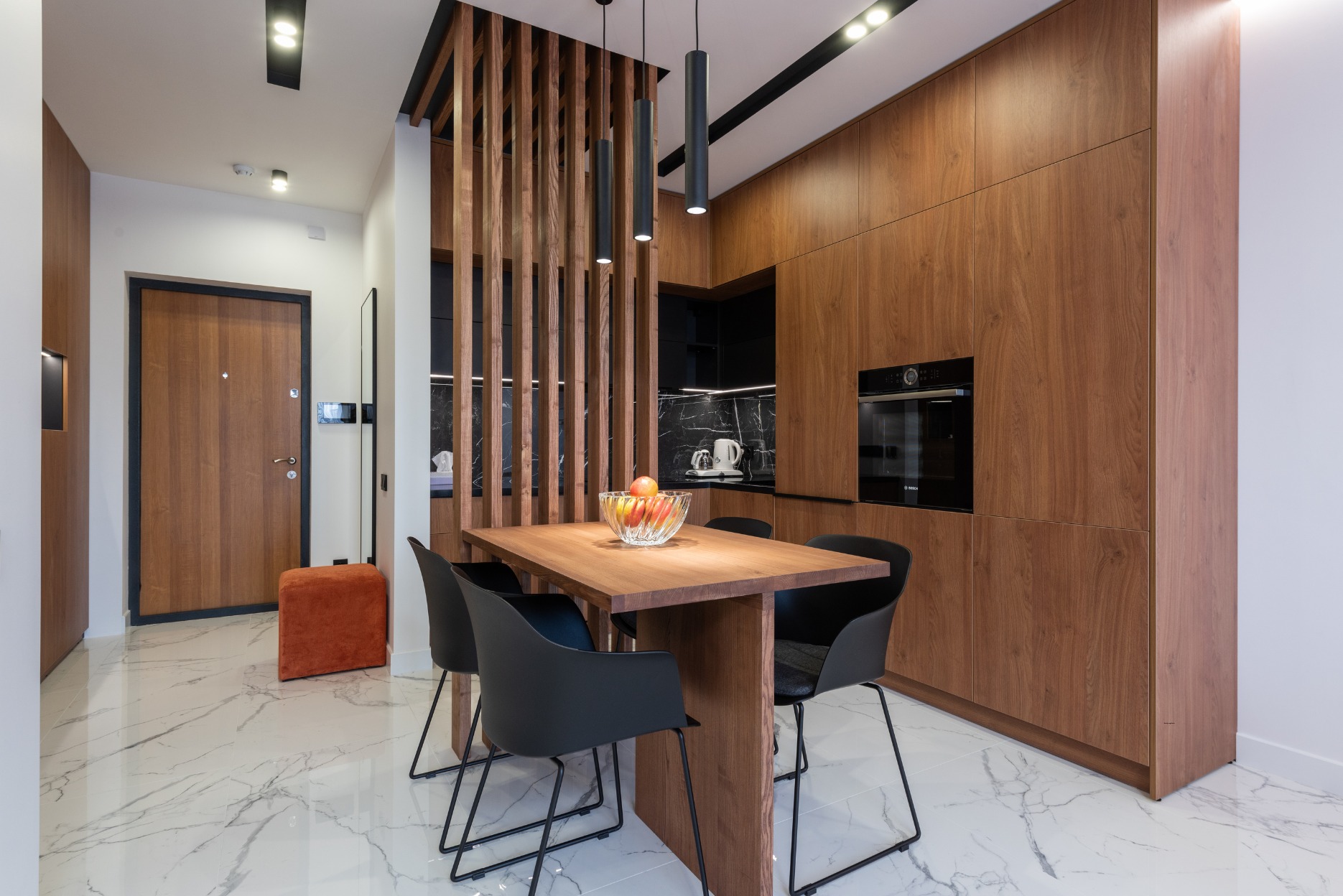 Nowoczesna kuchnia z drewnianymi szafkami, czarnym blatem i marmurową podłogą. Stół z owocami i czarne krzesła dodają elegancji.