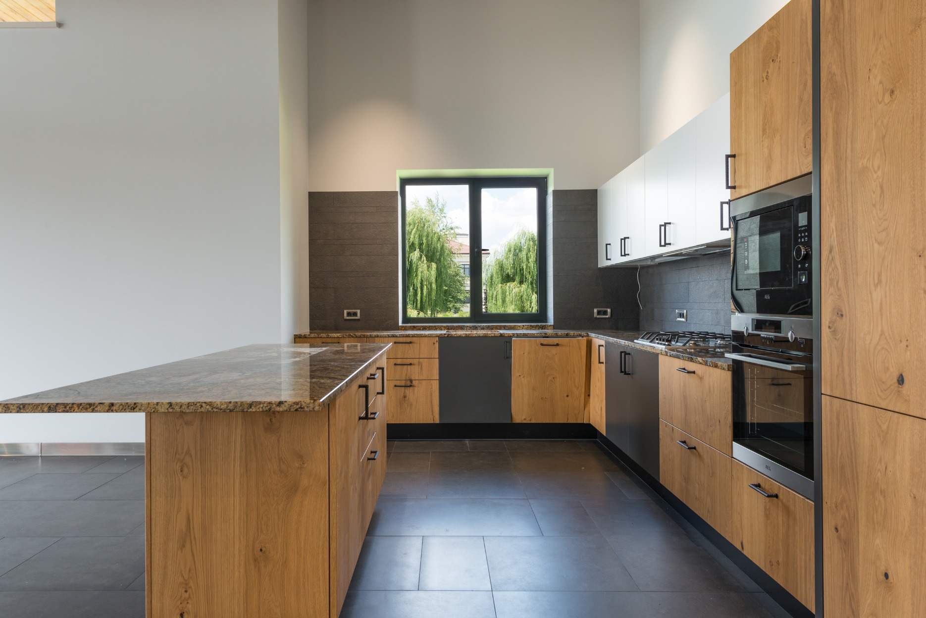 Nowoczesna kuchnia z drewnianymi frontami, czarnymi uchwytami i blatem z granitu. Okno z widokiem na zieleń.
