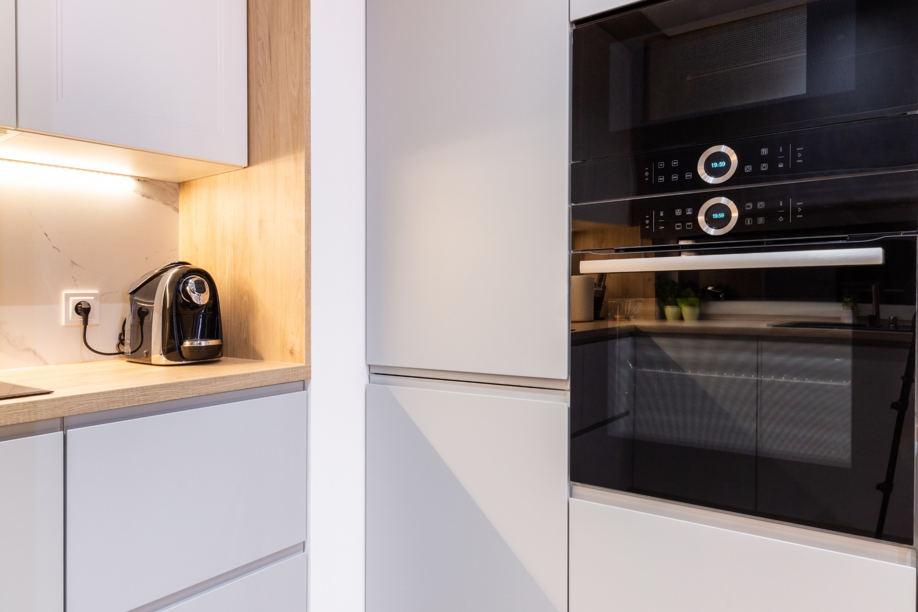 Nowoczesna kuchnia: zabudowa z szarymi frontami, drewniany blat z ekspresem do kawy i wbudowany piekarnik z mikrofalówką. Minimalistyczny design, jasne oświetlenie.